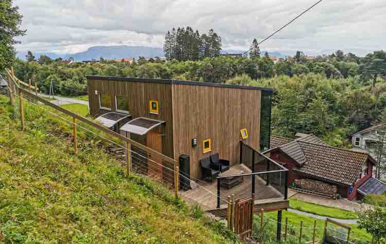A modern wooden house is positioned on a gentle slope, surrounded by greenery. Large windows with bright yellow frames stand out against the natural wood exterior. A spacious deck is visible, featuring outdoor seating and views of the scenic landscape.