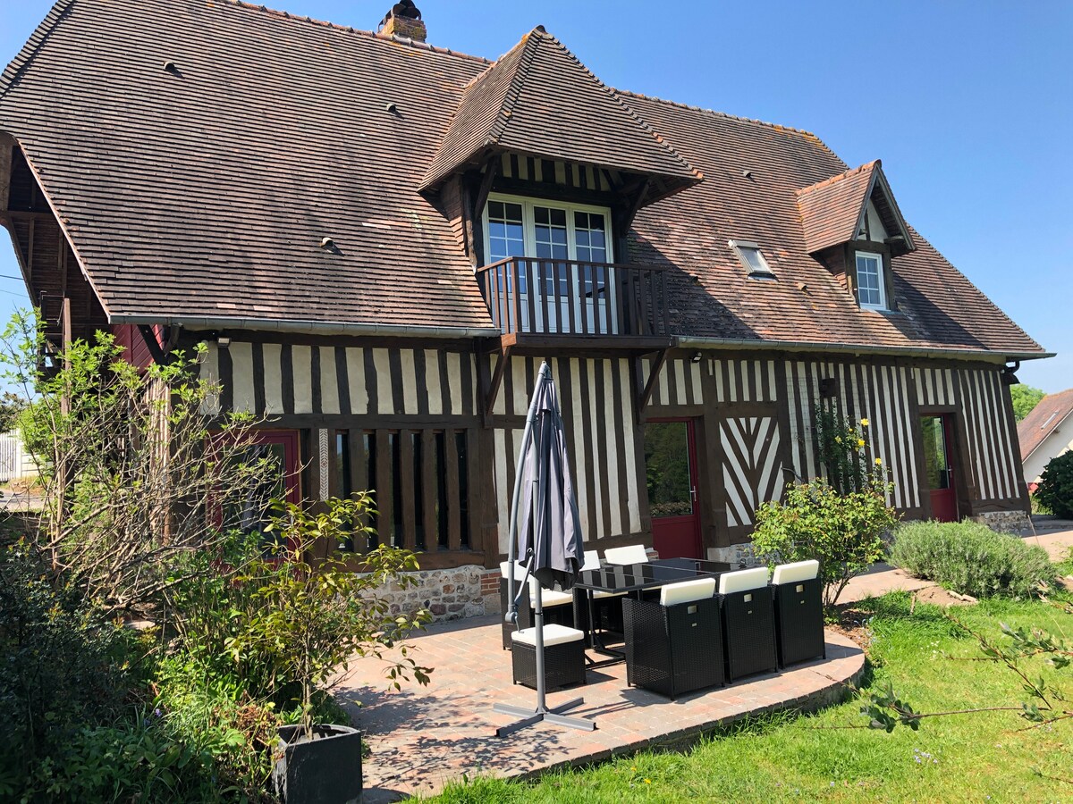The charming exterior of a restored Norman press features traditional timber framing and a large patio area. Outdoor seating, including a table and chairs, provides ample space for relaxation. Lush greenery surrounds the home, with a balcony visible on the upper level, offering scenic views.