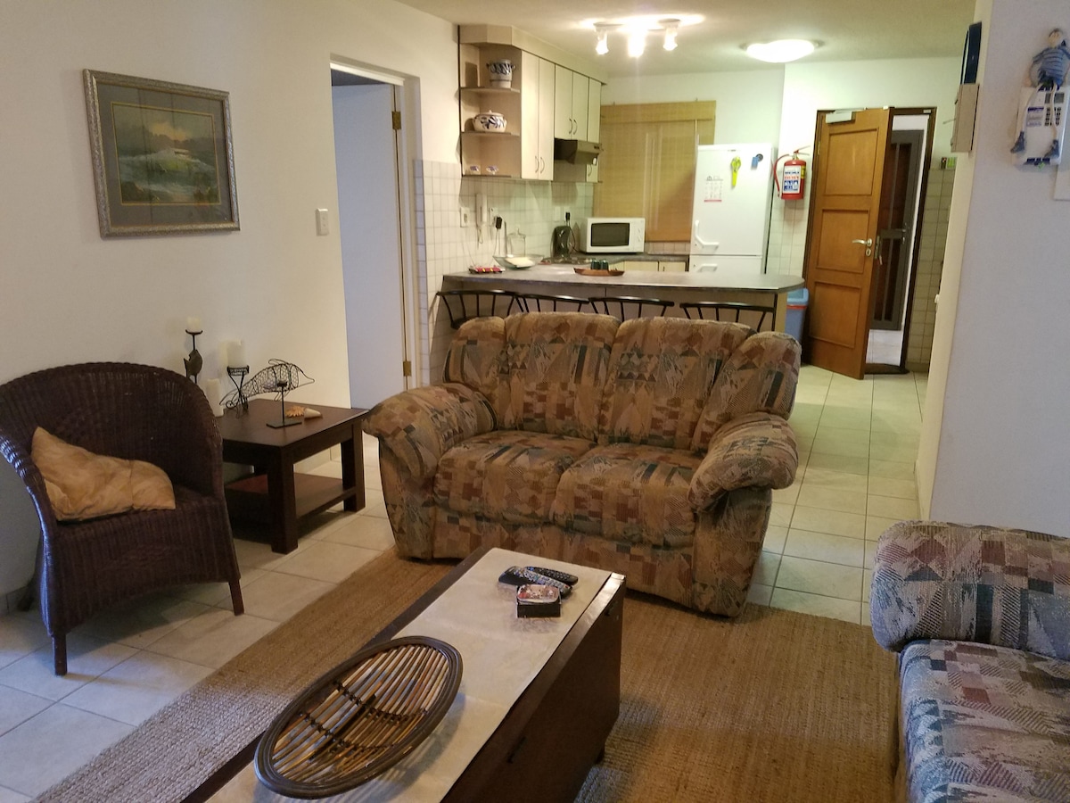 A living area is showcased with a patterned couch and a wicker chair. A coffee table is placed in front, and a textured rug is beneath. The space leads to a kitchen with cupboards and appliances visible in the background, creating an inviting environment for relaxation.