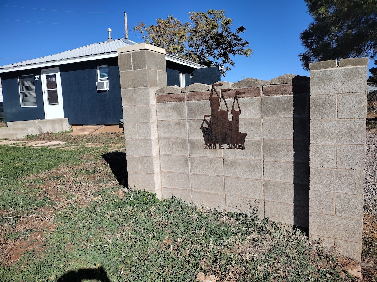 A sturdy stone wall is prominently featured, marked with the house number 265 E 300 S. The adjacent grassy area provides a natural contrast, while a blue house with a metal roof and air conditioning unit is visible in the background.