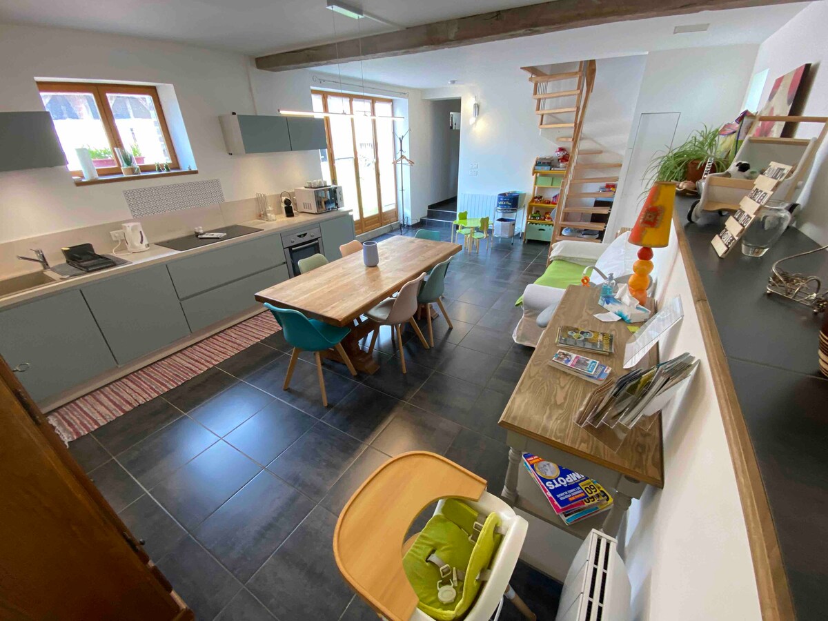 The kitchen and dining area feature modern cabinetry and appliances, accompanied by a spacious wooden dining table surrounded by colorful chairs. Natural light enters through large windows, illuminating the open space that connects to a cozy living area. A small bookshelf and children's toys are visible.