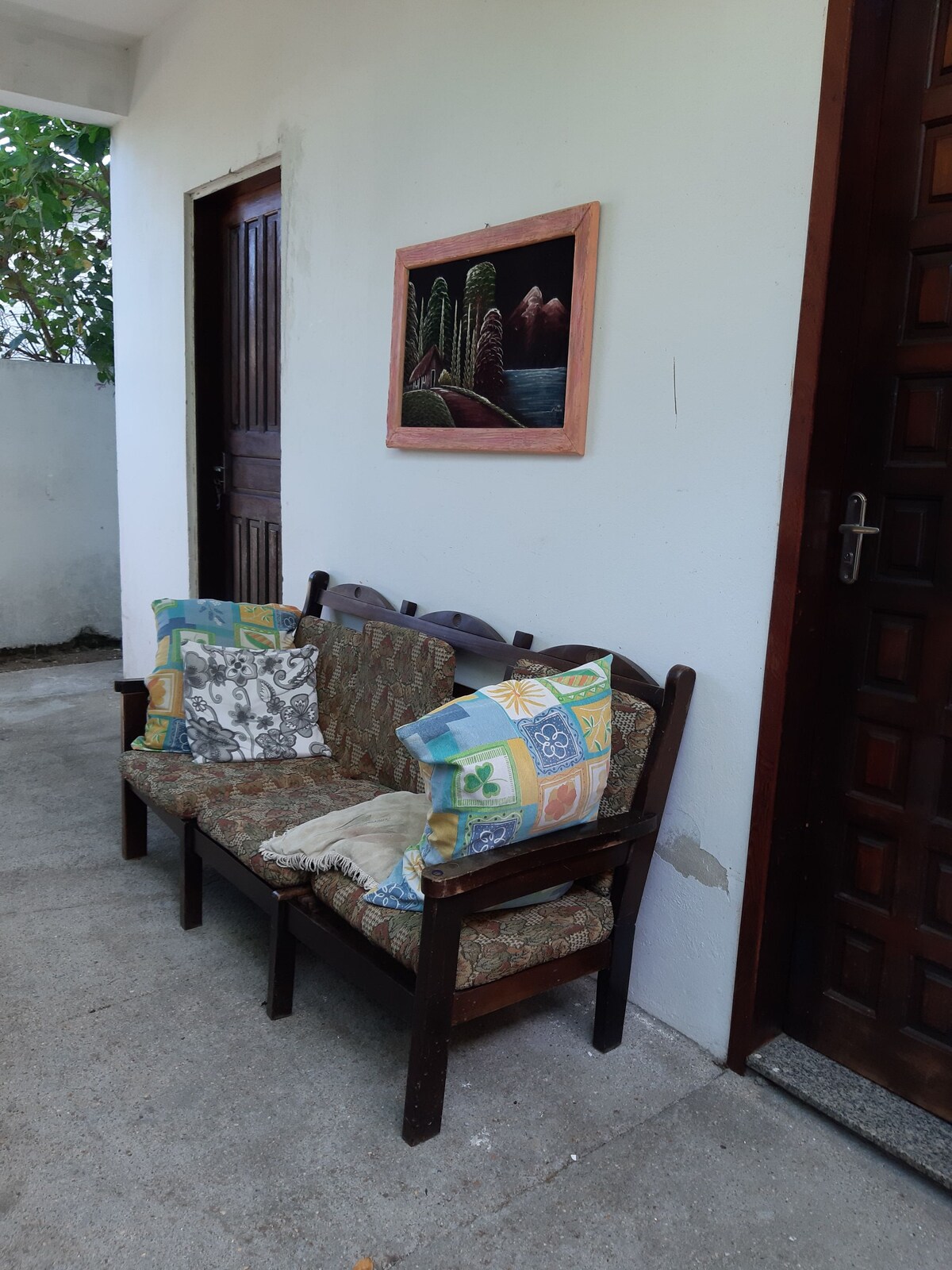 A simple outdoor seating area features a three-seat wooden bench adorned with patterned cushions. A framed artwork hangs on the wall nearby, and the entrance is marked by a dark wooden door, contributing to a welcoming environment.