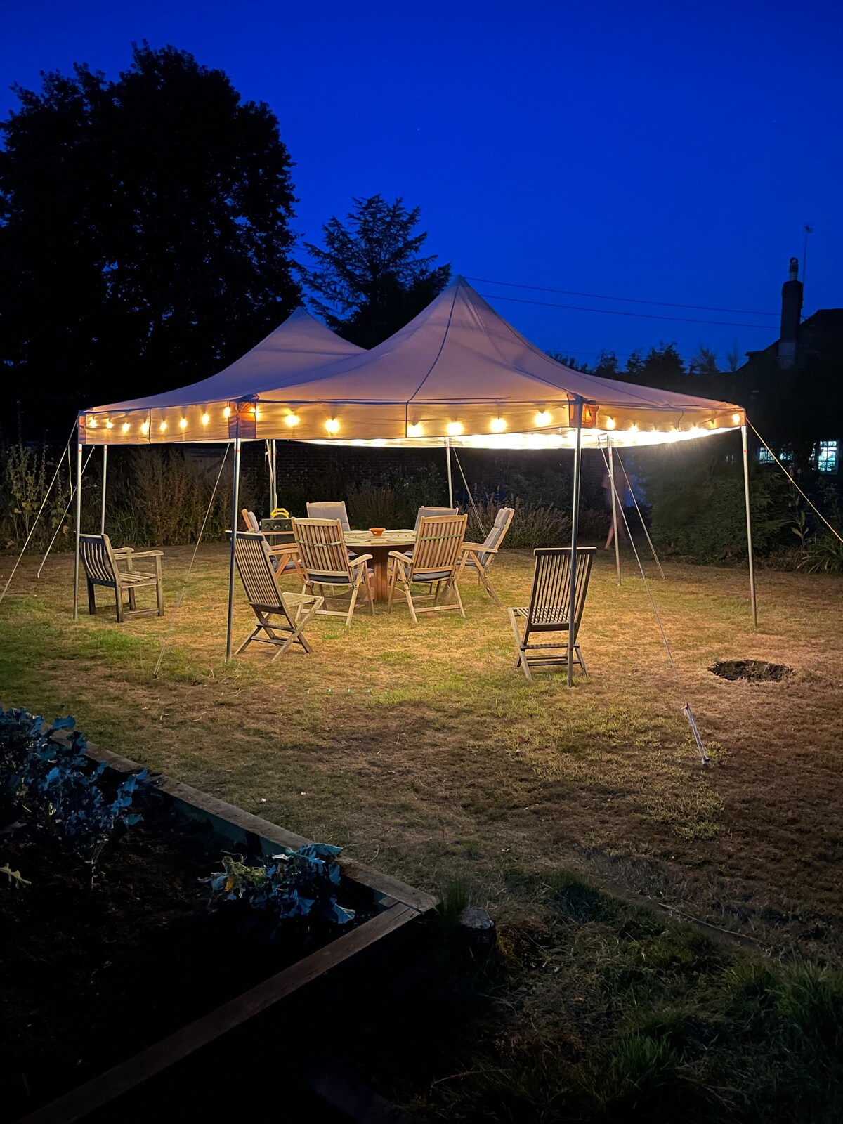 A marquee featuring warm string lights stands in a grassy area, surrounded by trees. Wooden garden furniture, including several chairs and a table, is arranged underneath, providing an inviting space for gatherings in the evening.