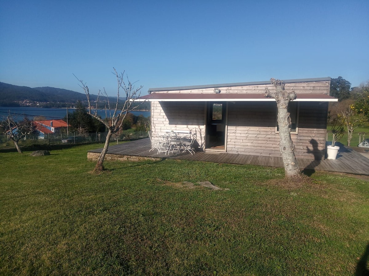 A wooden casita is depicted with large glass doors, surrounded by a lush green lawn. The structure faces the sea, providing expansive views. A wooden deck features a dining area with white chairs. Nearby trees and a peaceful landscape enhance the natural setting.