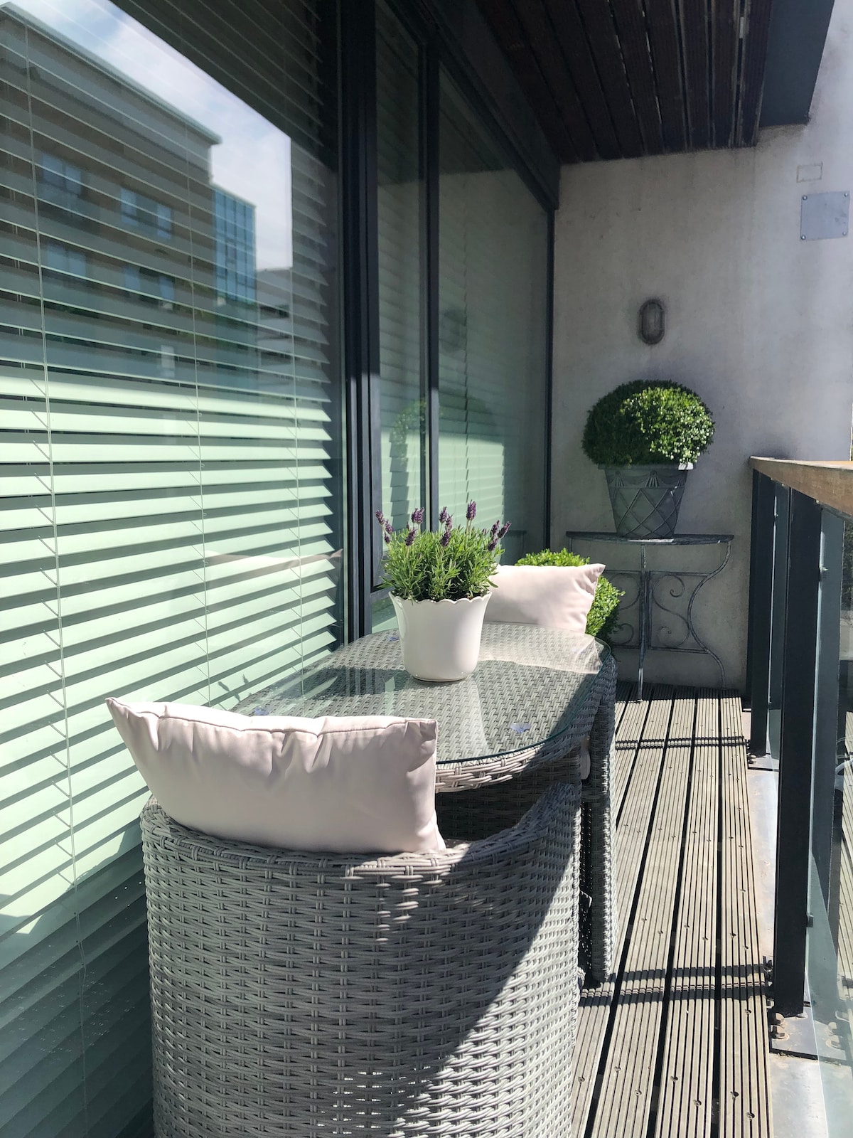 A private balcony is featured with a textured wooden floor, complemented by a small glass-top table and two woven chairs. Potted plants, including lavender and a rounded shrub, add greenery, while sunlight filters through the blinds, creating a serene outdoor space.