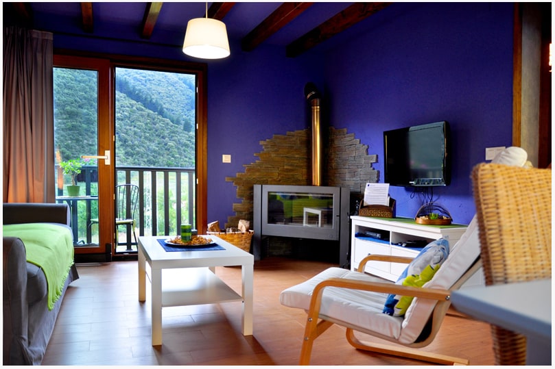 A modern living area features a vibrant purple wall complemented by wooden beams. A cozy seating arrangement includes a gray sofa and a white armchair. Natural light floods the space through large glass doors, revealing views of the surrounding greenery. A fireplace is positioned against a stone wall.