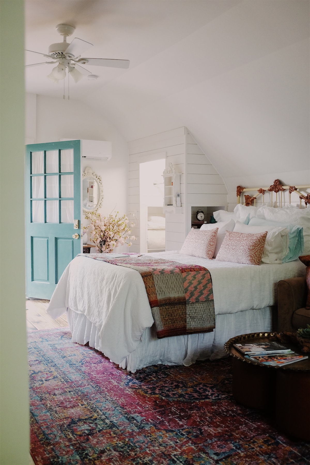 A cozy sleeping area is characterized by soft bedding layered with a decorative throw. A vibrant area rug adds color and warmth, while a ceiling fan and air conditioning unit ensure comfort. A charming blue door leads to the entrance, inviting natural light into the space.