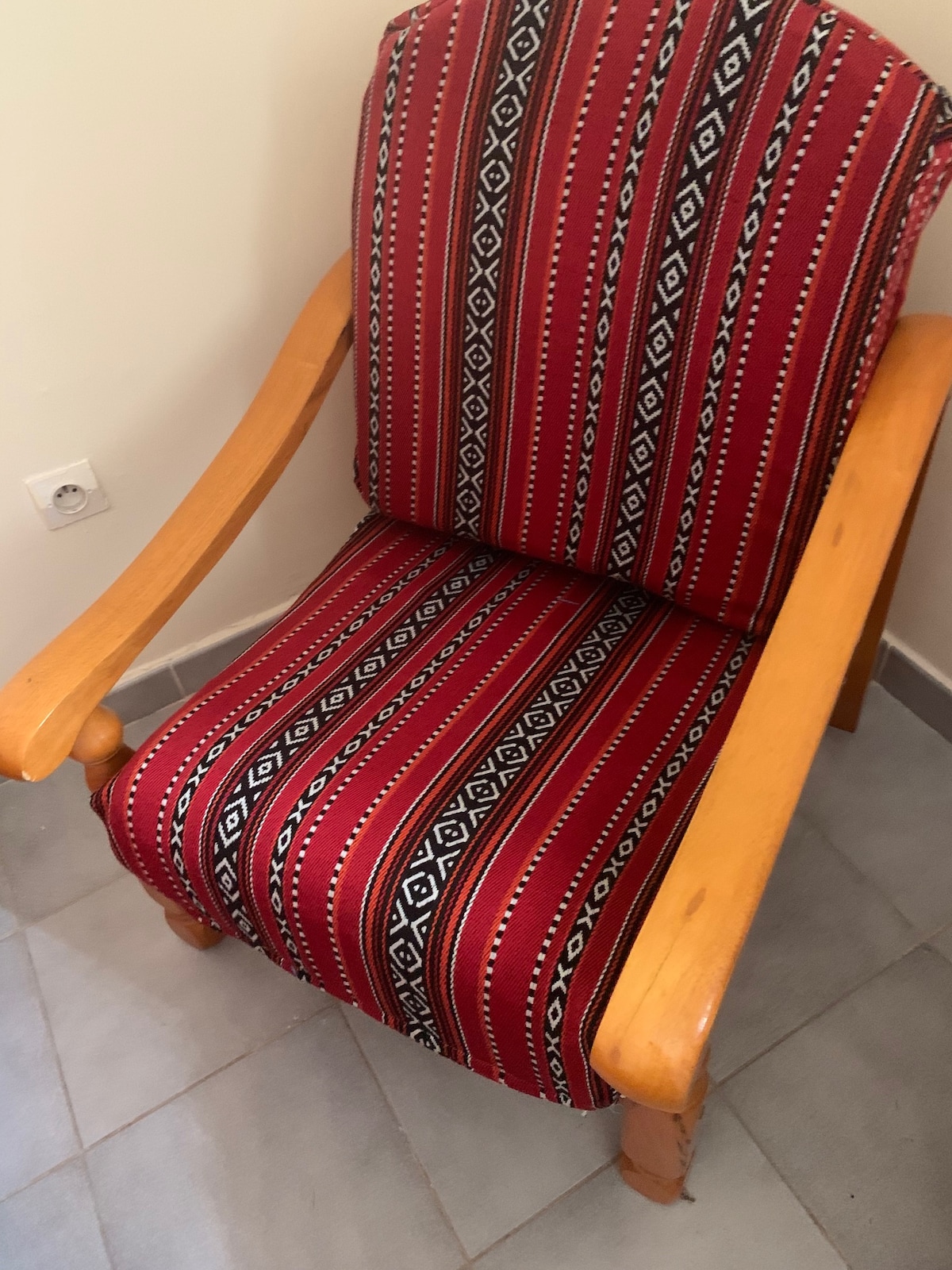 A traditional chair with a wooden frame is adorned with red and black fabric featuring geometric patterns. The chair is positioned on a tiled floor, contributing to a cozy seating area.