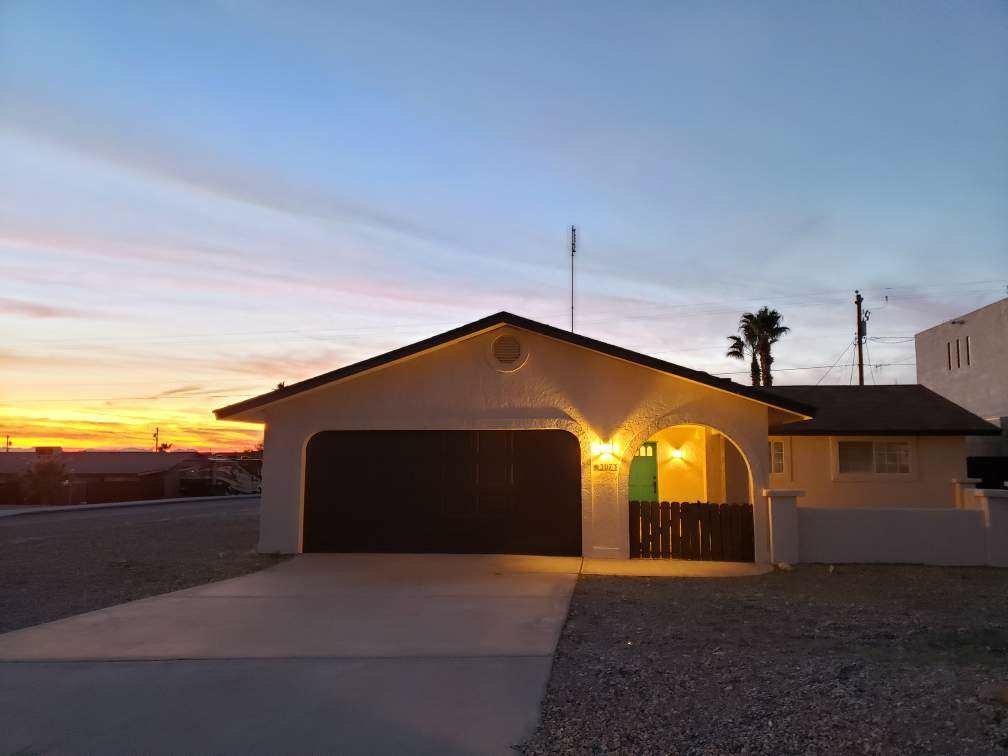 A single-story home is depicted with a warm glow emanating from the garage side. A driveway leads up to the structure, while a serene sunset paints the sky in soft pastel colors. The presence of palm trees adds to the outdoor ambiance.