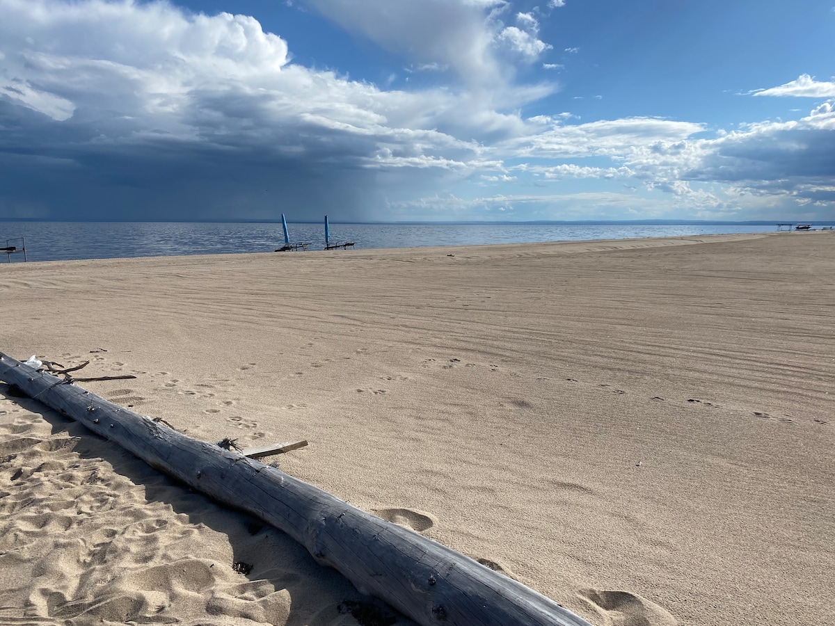 An expansive sandy beach stretches towards the horizon, with gentle waves lapping at the shore. A weathered log lies in the foreground, while distant blue structures are visible on the water's edge, under a sky filled with clouds that cast soft shadows.