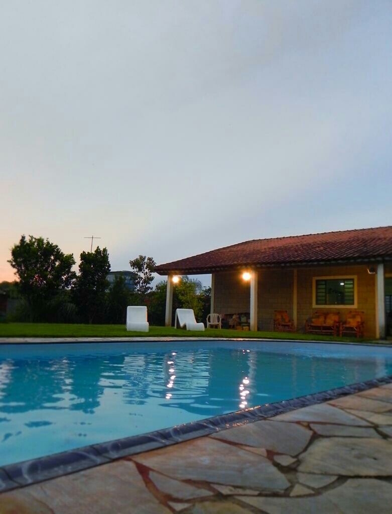 A serene pool area features a clear water pool surrounded by smooth stone decking. Two reclining lounge chairs are positioned by the water, while a covered seating area with wooden furniture can be seen beside the house. Lush greenery and a soft sky complete the scene.