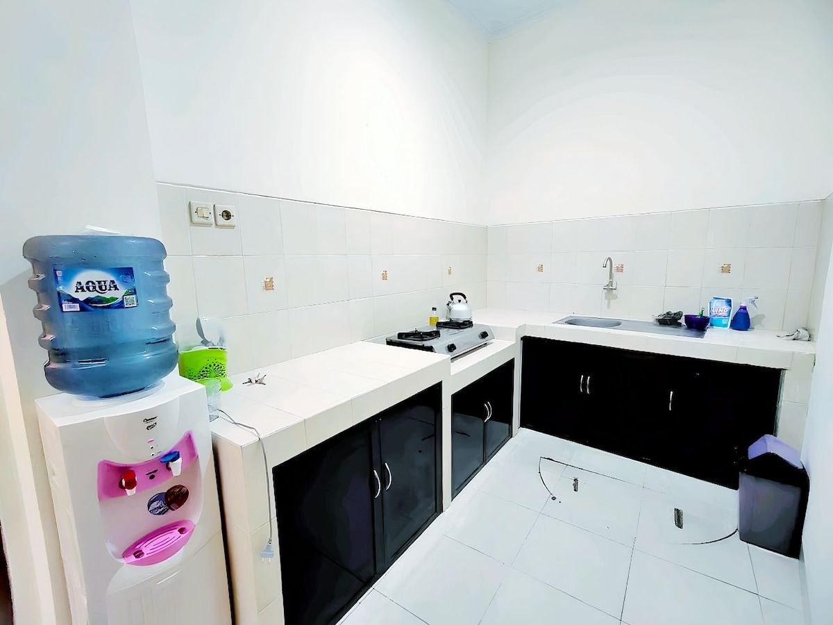 A clean kitchen area is equipped with dark cabinetry and white countertops. A water dispenser is positioned to the left, with a kettle placed on the stove. A sink is located at the back, and cleaning supplies are arranged neatly on the countertop.