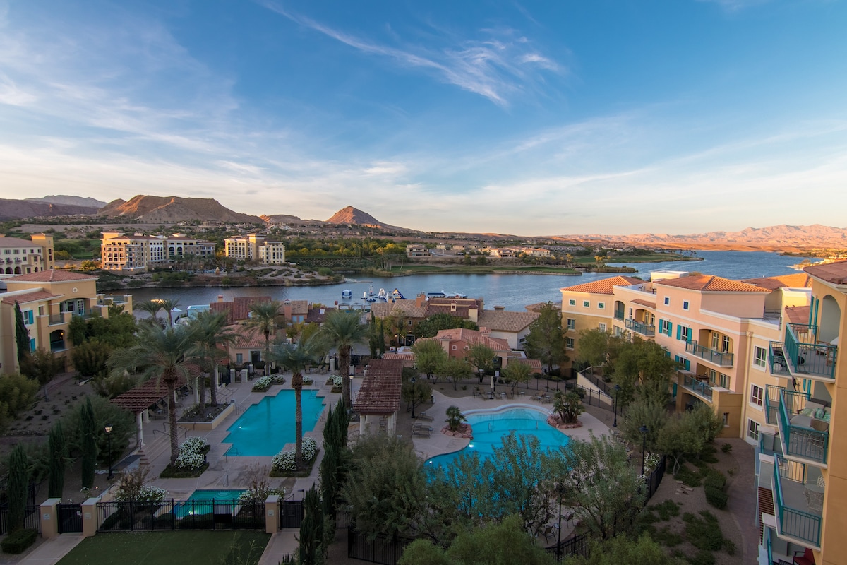 A panoramic view showcases Lake Las Vegas with its glistening water and surrounding mountains. Two inviting pools are visible in the foreground, framed by swaying palm trees and lush landscaping, while elegant Mediterranean-style buildings enhance the serene lakeside atmosphere.