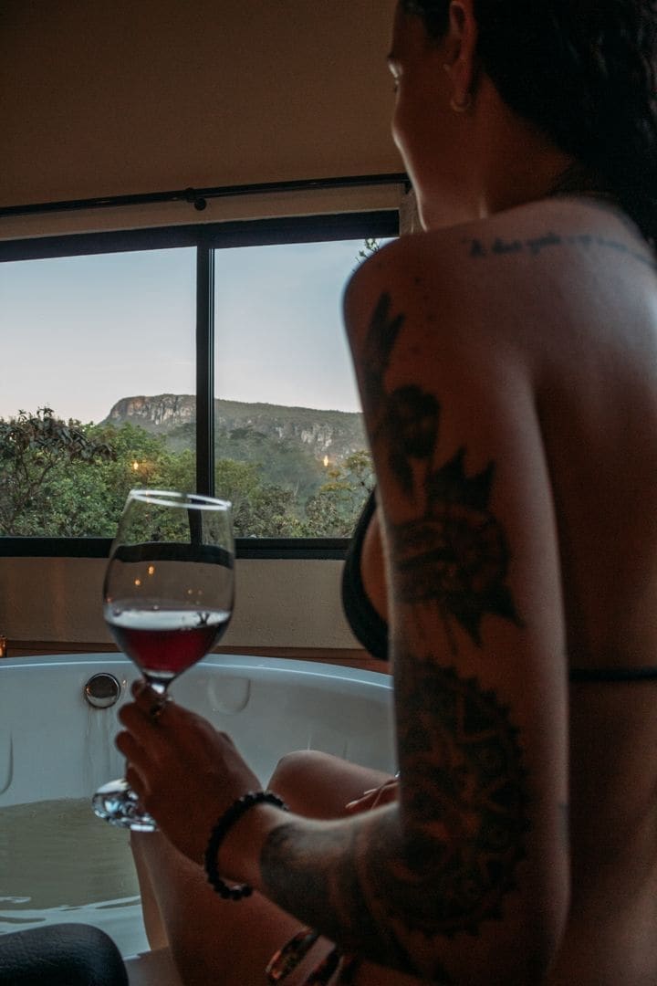A scenic bathroom features a large soaking tub positioned near a window, allowing views of distant mountains. A glass of red wine is held beside the tub, adding to the relaxing ambiance of the space.