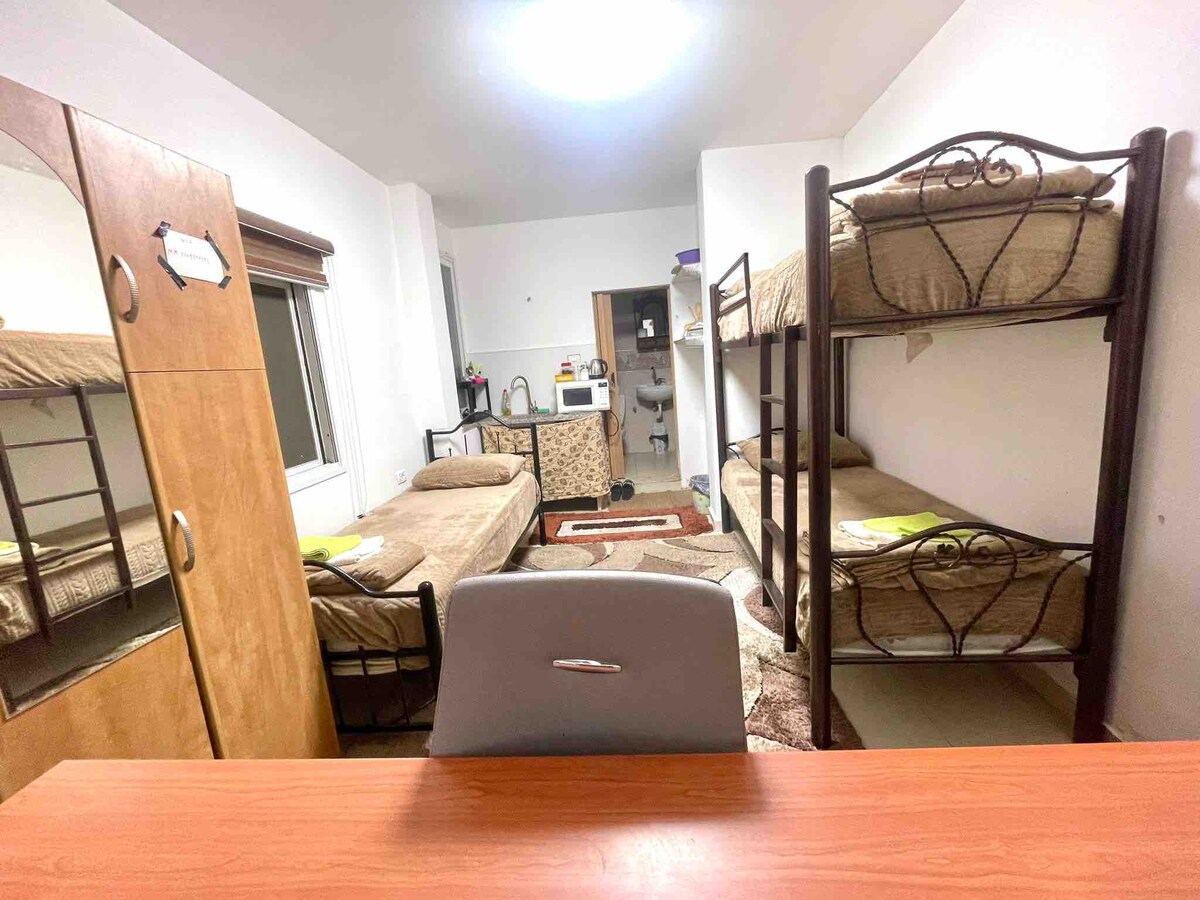 A comfortable room features two sets of bunk beds with soft bedding and a workspace area with a desk and chair. A small kitchenette is visible, equipped with essential appliances. Natural light enters through a window, enhancing the simple and functional layout.
