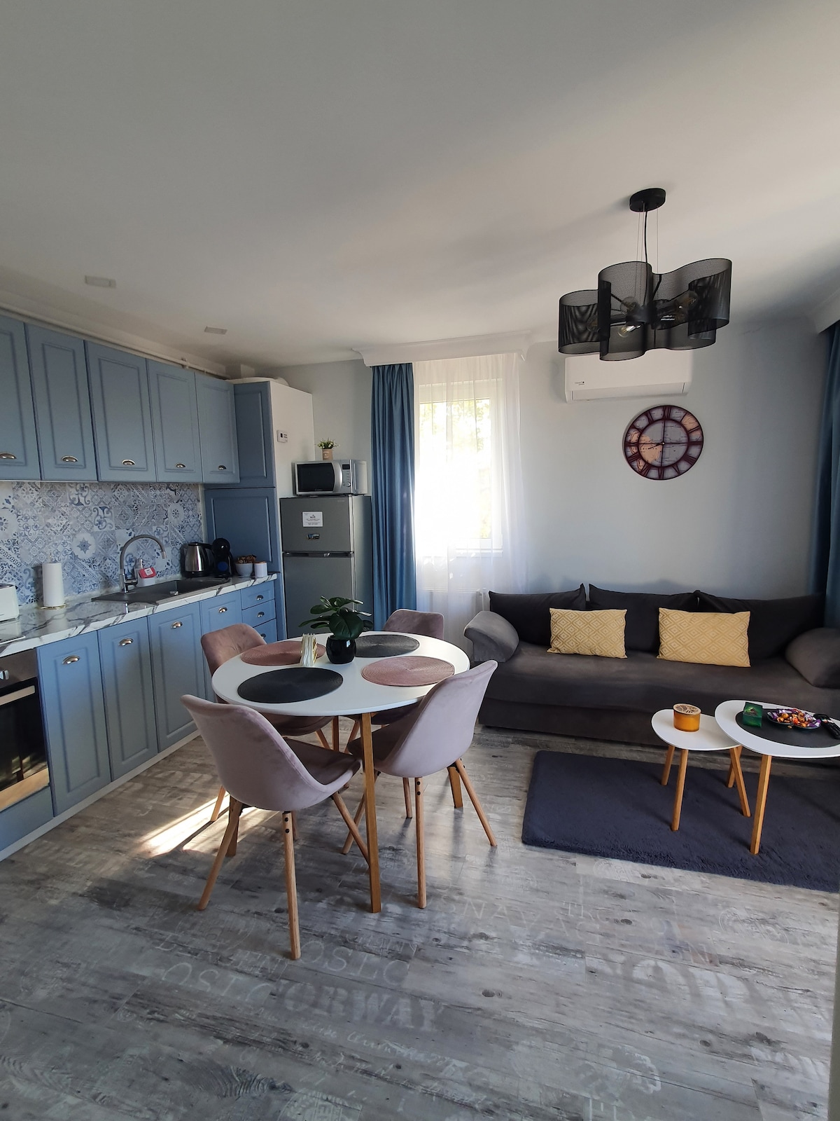 The open space kitchen and living area feature light blue cabinetry and modern appliances. A round dining table with four chairs is placed beside a comfortable sofa adorned with cushions. A large clock and decorative bowl with snacks are also visible, enhancing the inviting setting.