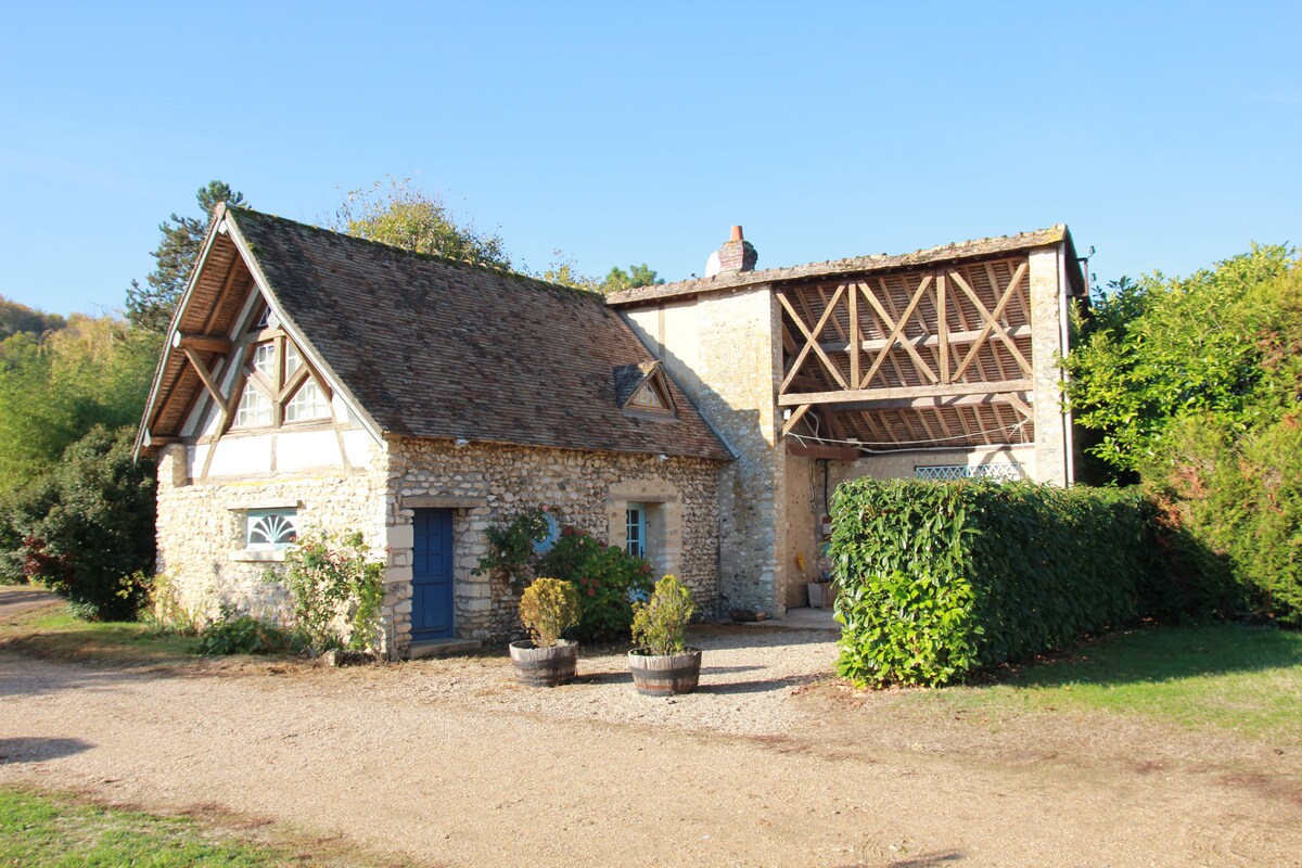 The exterior of an old farmhouse showcases a stone and timber structure under a clear blue sky. The entrance is adorned with blue doors, while lush greenery encircles the building, offering a sense of tranquility and connection to nature.