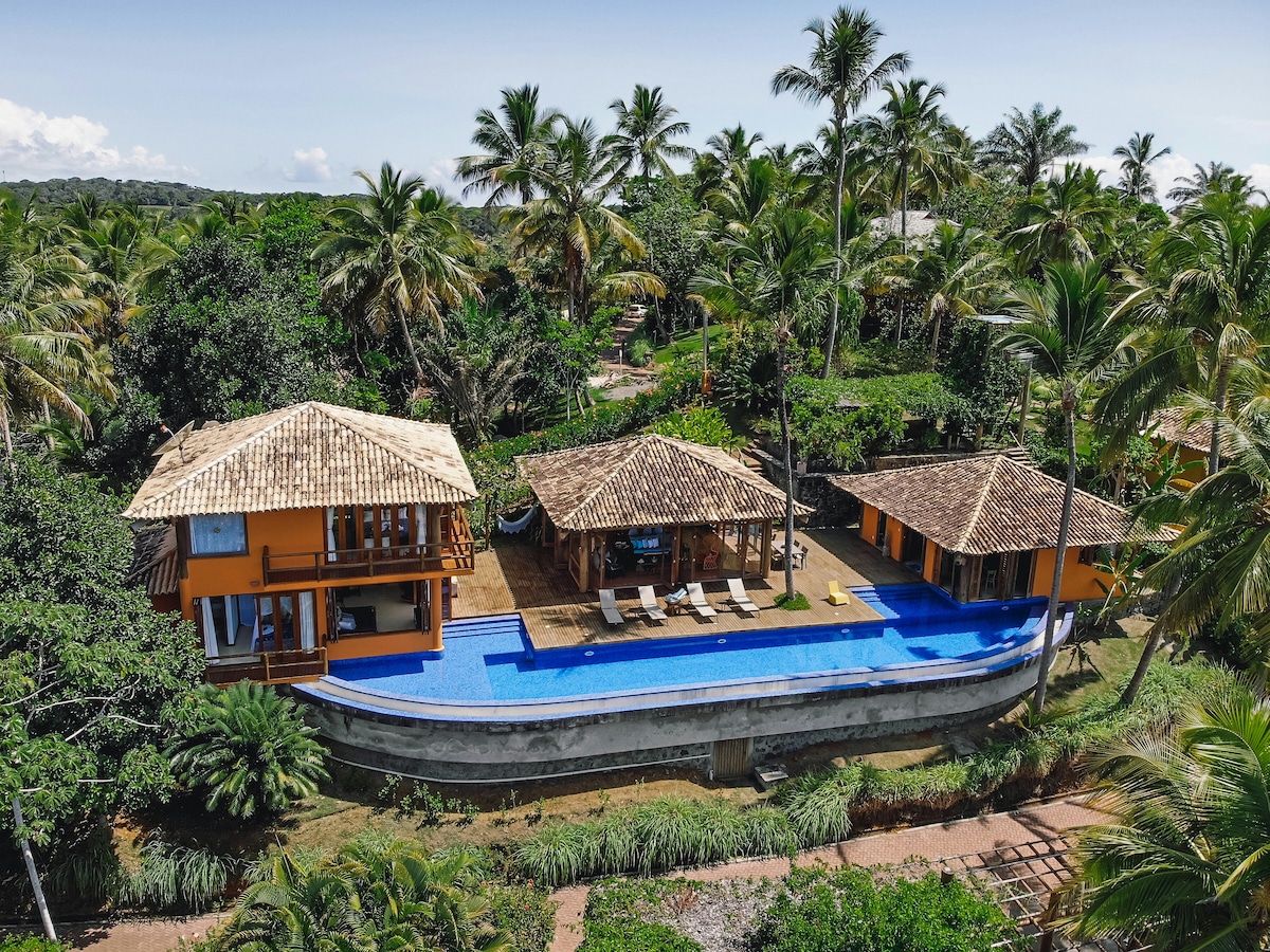 An aerial view captures the villa with its distinctive architecture surrounded by lush greenery. A long blue pool stretches between the structures, complemented by palm trees. The roofs feature natural materials, and spacious decks are visible, providing open areas for relaxation.