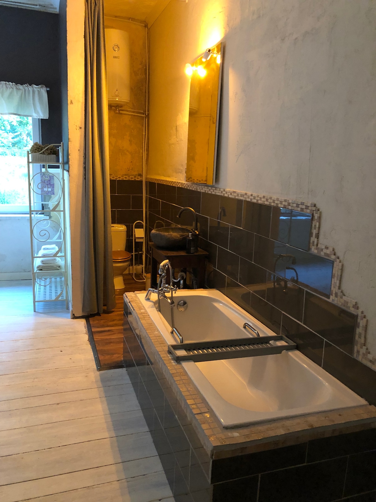 An integrated bathroom features a spacious bathtub with fixtures and a wooden shelf above. Warm lighting accentuates the tiled wall and reflects off the marble counter. A partially draped curtain offers privacy while a glimpse of natural light enters from a nearby window.