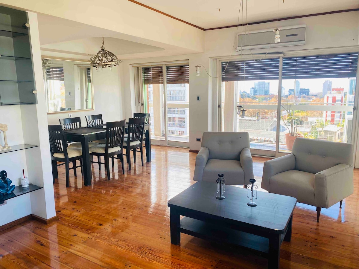 A bright living and dining area features large windows with city views. A dark wooden coffee table sits between two light gray chairs. A dining table with black chairs accommodates six people. Natural light fills the space, enhancing the polished wooden floors.