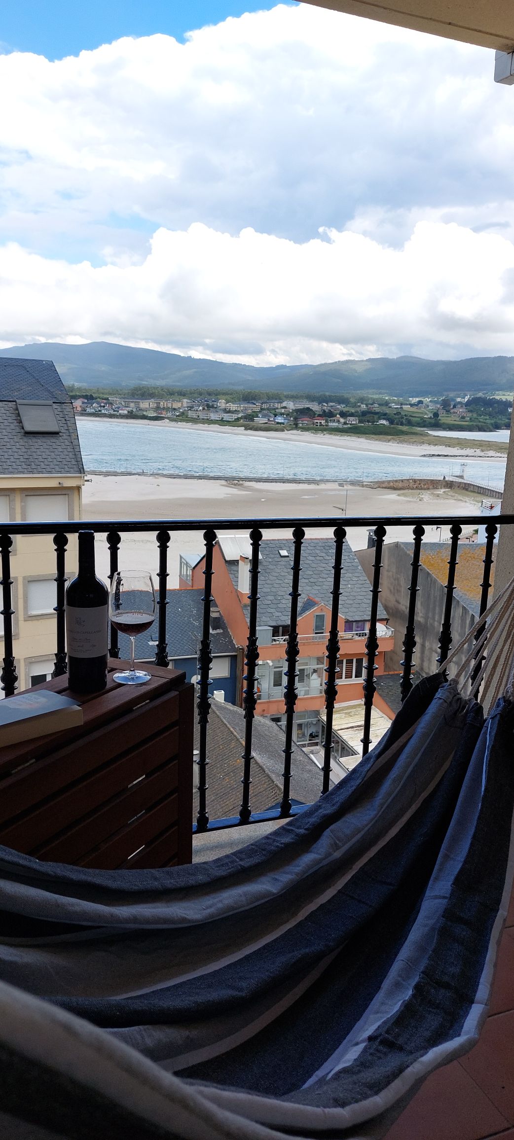 A balcony features a comfortable hammock, complemented by a small wooden table holding a wine bottle and glass. The view encompasses a calm body of water, with distant mountains and colorful buildings lining the shoreline.