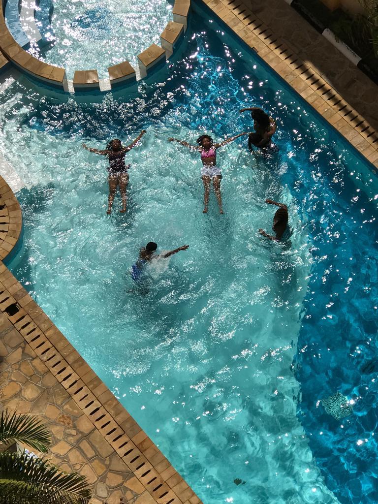 The swimming pool is surrounded by a stone deck, featuring clear blue water that glitter under the sunlight. Several individuals are seen enjoying the water, with some floating and others playing, creating a vibrant atmosphere on a warm day.
