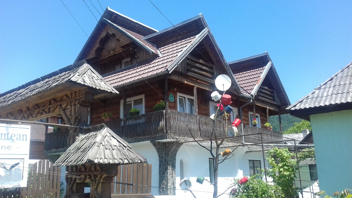 A wooden house with distinctive architecture features multiple balconies adorned with flower pots. The sloped roof with wooden shingles adds to the rustic charm. A nearby structure with a thatched roof completes the picturesque setting against a clear blue sky.