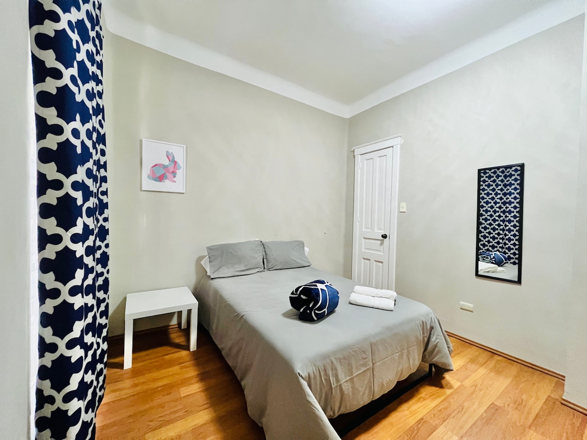 A simple bedroom is furnished with a spacious bed covered in gray linens. A mirrored wall reflects natural light, enhancing the room's brightness. A small white side table accentuates the space, while rolled towels are neatly arranged at the foot of the bed.