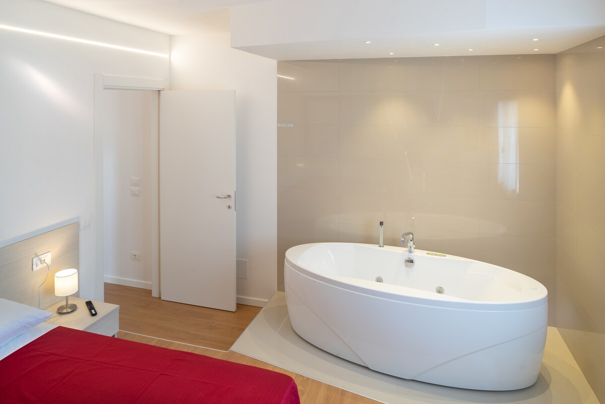 A modern room features a spacious oval bathtub positioned near the wall. Soft ambient lighting enhances the serene atmosphere. A neatly made bed is visible with red bedding, and a subtle door leads to another area of the apartment.