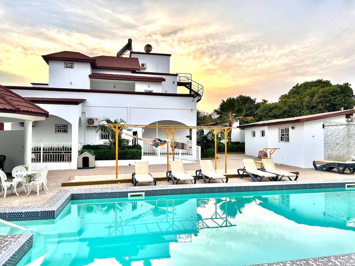 A tranquil swimming pool, reflecting the sunset, is surrounded by lounge chairs and a shaded seating area. The property's white facade is visible in the background, bordered by lush greenery, creating a serene outdoor space for relaxation.