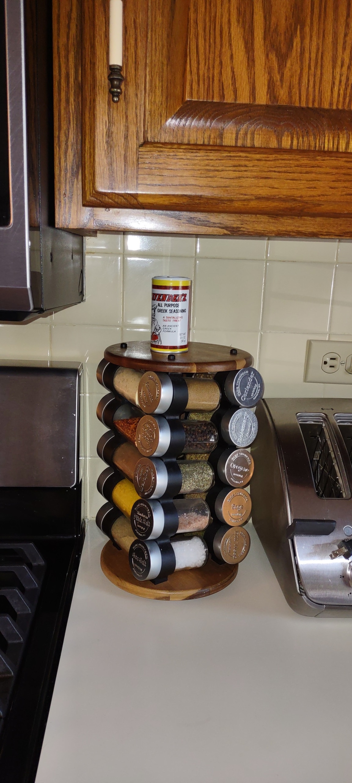 A tiered spice rack is displayed on a countertop, featuring various containers of spices and herbs organized in a circular arrangement. The dark containers contrast with the light wood of the rack, and a bottle of seasoning is placed on top. Kitchen appliances, including a toaster, are partially visible in the background.