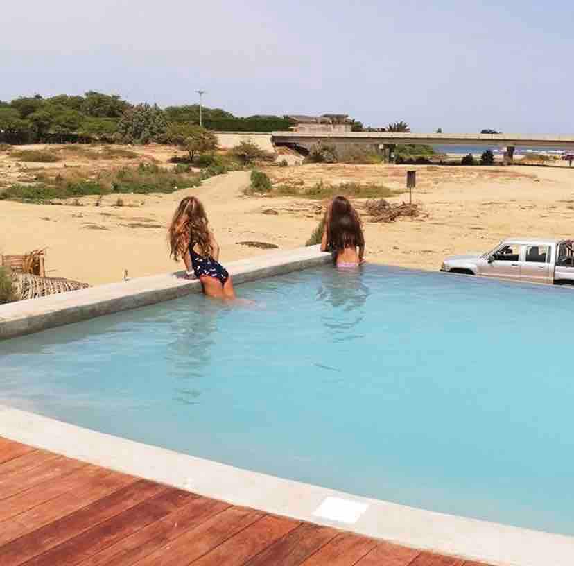 An infinity pool extends towards the horizon, with two individuals enjoying the water. Sunlit sand dunes and greenery are visible in the background, along with a distant vehicle. The serene setting offers a peaceful atmosphere for relaxation.