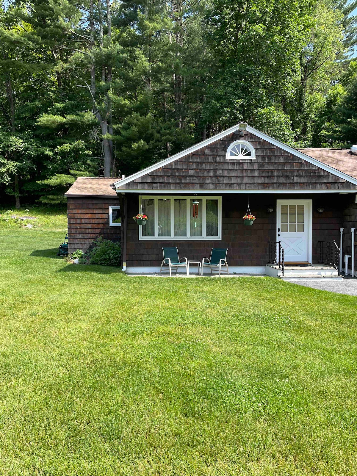 A charming exterior of a studio is shown, featuring a brown shingle façade and a welcoming front door. Two chairs are positioned on the grassy lawn, surrounded by lush greenery and tall trees. Flower baskets add a touch of color near the window.