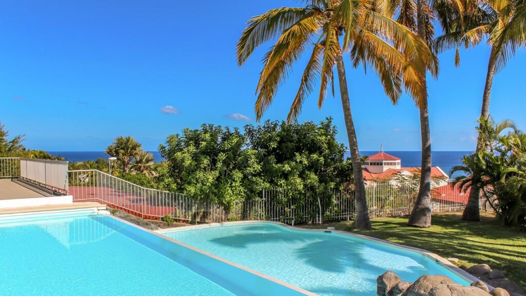 A clear view of a shared swimming pool is shown, flanked by palm trees and lush vegetation. The ocean can be seen in the distance under a bright blue sky, creating a serene outdoor space for relaxation.