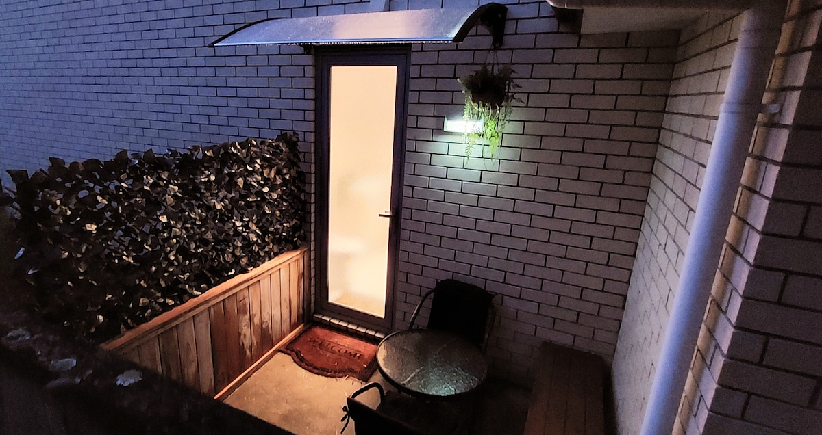 An outdoor patio area features a small round table accompanied by a chair. The space is enclosed by a low wooden fence with greenery. A door with a frosted window provides access, and a light illuminates the area, enhancing the evening ambiance.