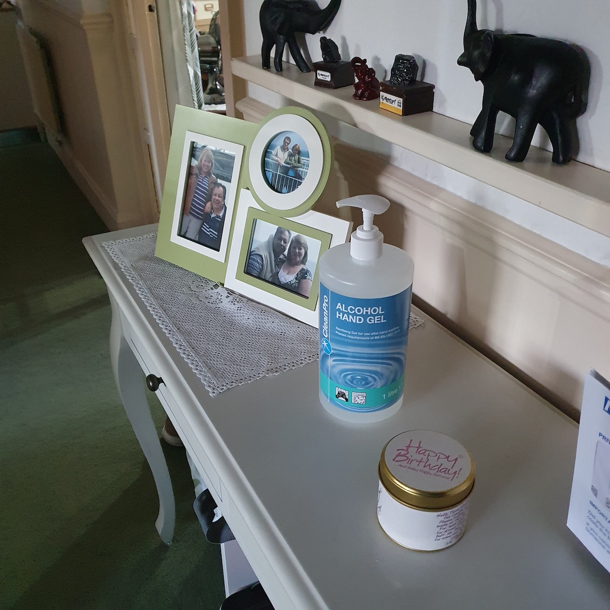 A console table is displayed with framed photographs and decorative items. An alcohol hand gel and a small jar are positioned on the surface, offering practicality. The elegant table features a lace runner, complementing the clean lines of the surrounding decor.
