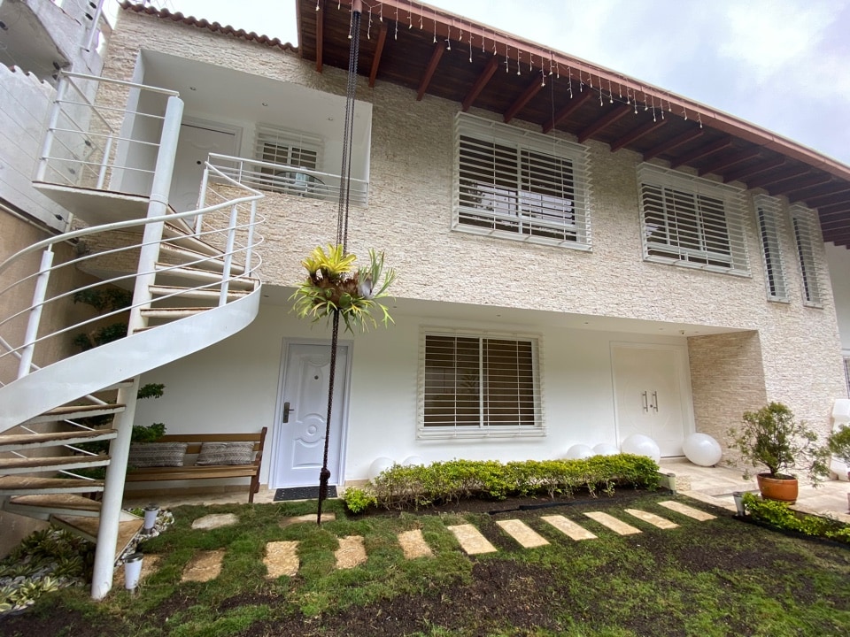 An exterior view of the loft-style suite reveals a light-colored building with a decorative stone facade. A spiral staircase leads to the upper level. Flanking the entrance, well-maintained green areas feature neatly arranged stones, while large windows provide ample natural light.