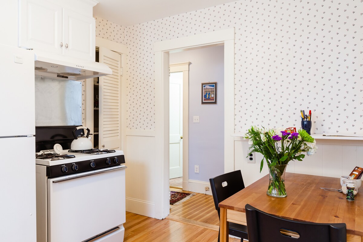 A clean kitchen space is visible, featuring a white gas stove and refrigerator. A wooden dining table is set with black chairs and a vase of fresh flowers. A doorway leads into a light-colored hallway, enhancing the inviting atmosphere of the area.