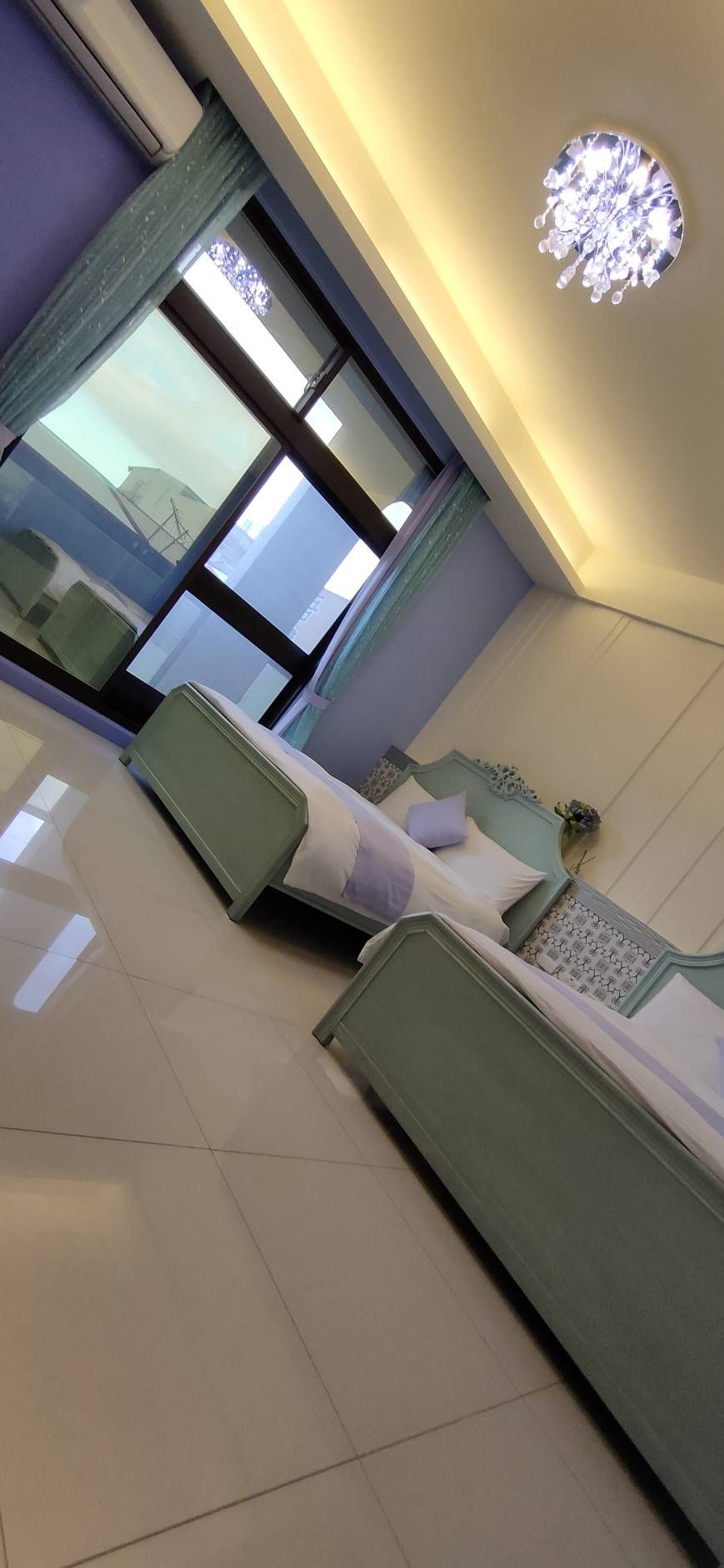 A bright and airy guest room is presented with two twin beds featuring soft blue accents. Large glass windows offer natural light and a view of the outside, while a stylish chandelier adds a touch of elegance to the space.
