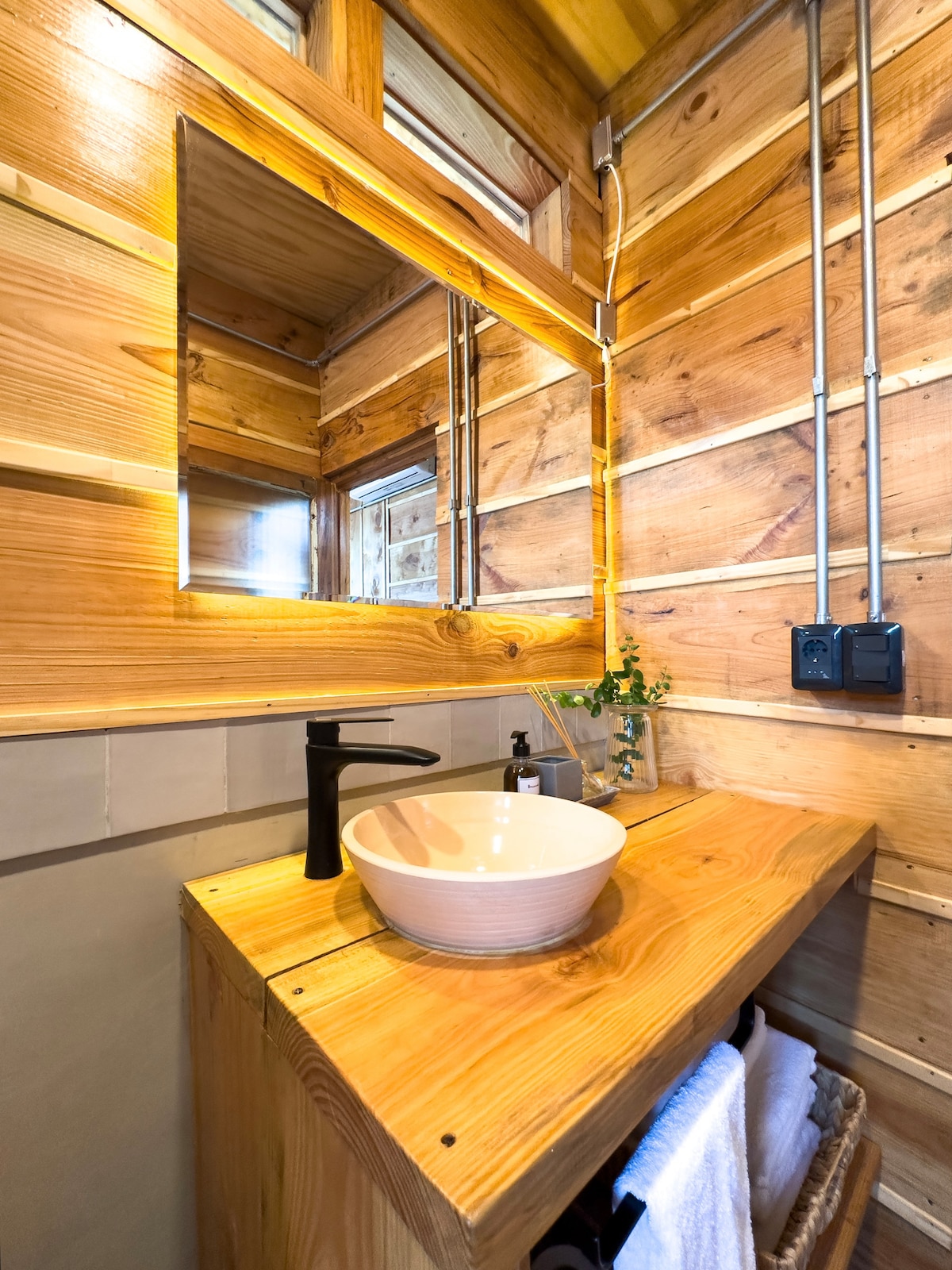 A wooden bathroom is featured, with natural wood paneling on the walls. A simple bowl sink rests on a wooden countertop, accompanied by a black faucet. A mirror is mounted above the sink, reflecting natural light from a nearby window. Fresh greenery adds a touch of nature.