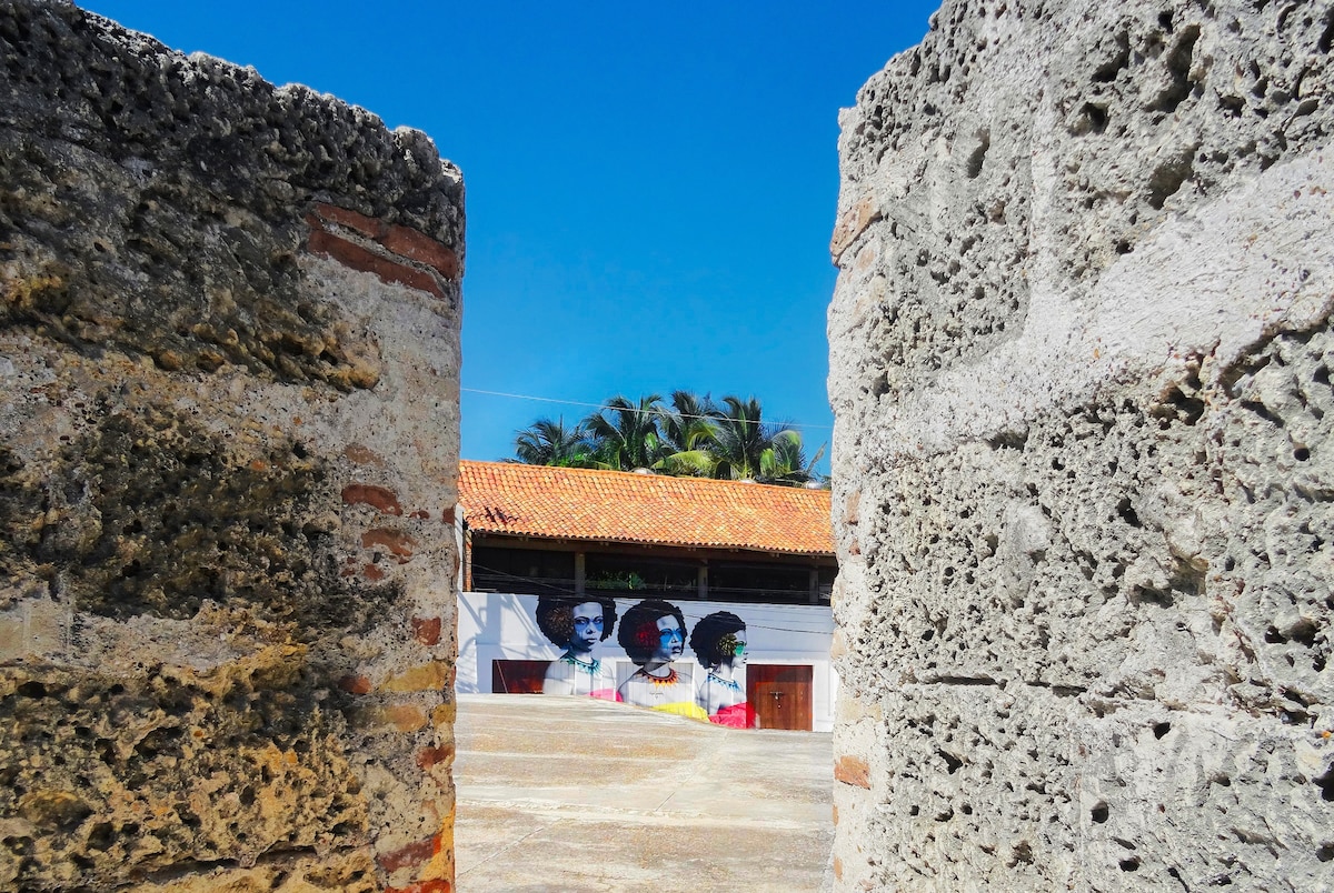 A clear view is provided through the stone walls, framing a vibrant mural depicting faces against a backdrop of a sunny blue sky. The distinct architectural features of the neighboring building and surrounding palm trees are also visible.