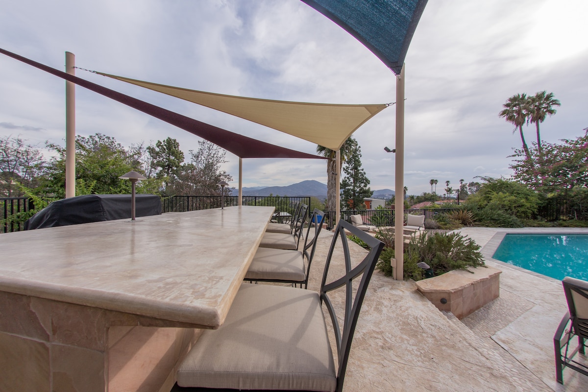 A spacious outdoor dining area features a long stone table surrounded by multiple cushioned chairs, shaded by colorful sail canopies. A beautiful pool and surrounding greenery are visible in the background, complemented by mountain views and palm trees under a cloudy sky.