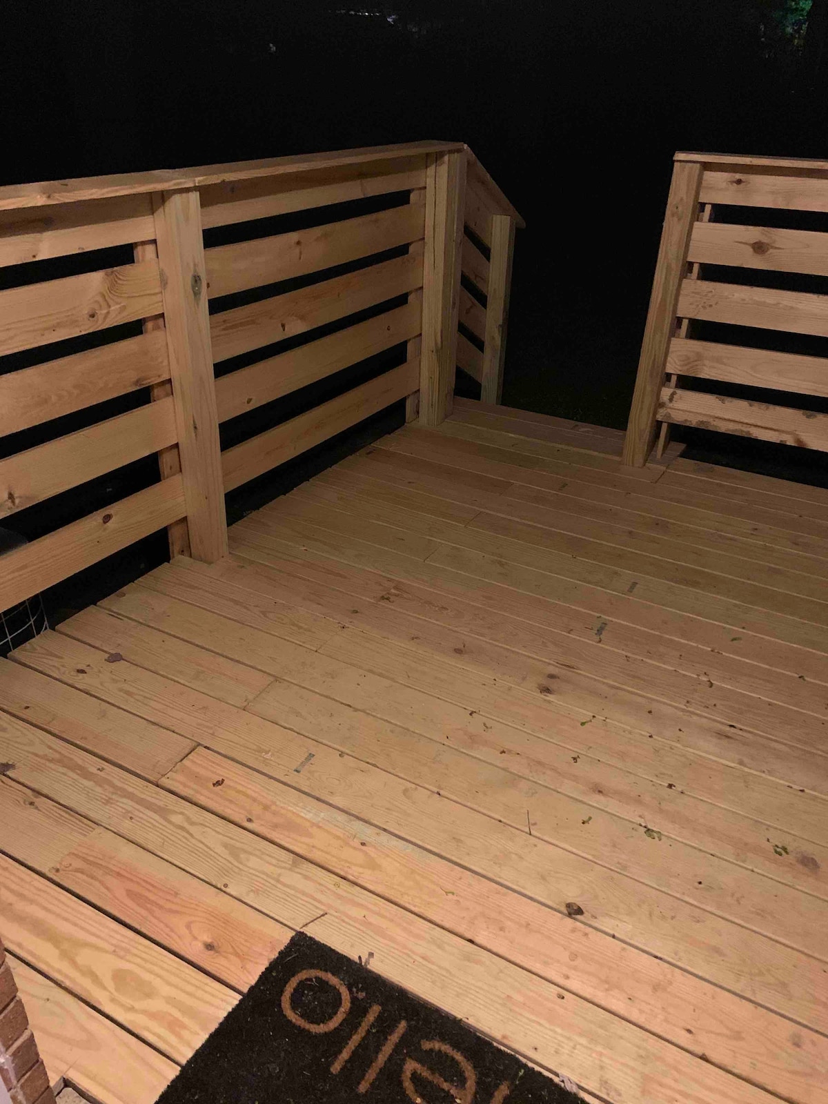 A wooden porch is displayed, featuring a sturdy railing and a slatted design. The planked flooring shows a natural wood finish, and a welcome mat is positioned near the entrance. The surrounding area appears dark, suggesting a serene evening setting.