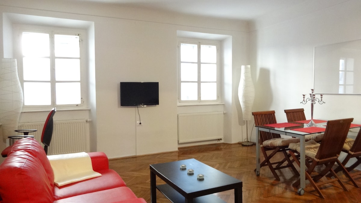 A spacious living area features large windows allowing natural light, enhancing the bright atmosphere. A red sofa is positioned comfortably next to a coffee table. A dining table with wooden chairs is set nearby, complemented by soft lighting from floor lamps.
