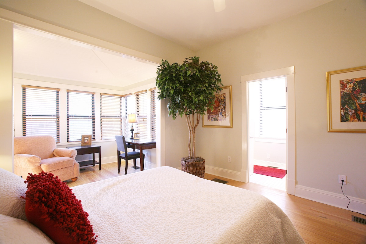 A bright bedroom showcases a spacious bed with a textured white coverlet, complemented by a decorative red pillow. Sunlight pours through multiple windows adorned with wooden blinds. A comfortable armchair and small desk are visible in the adjoining area, enhancing the inviting atmosphere.