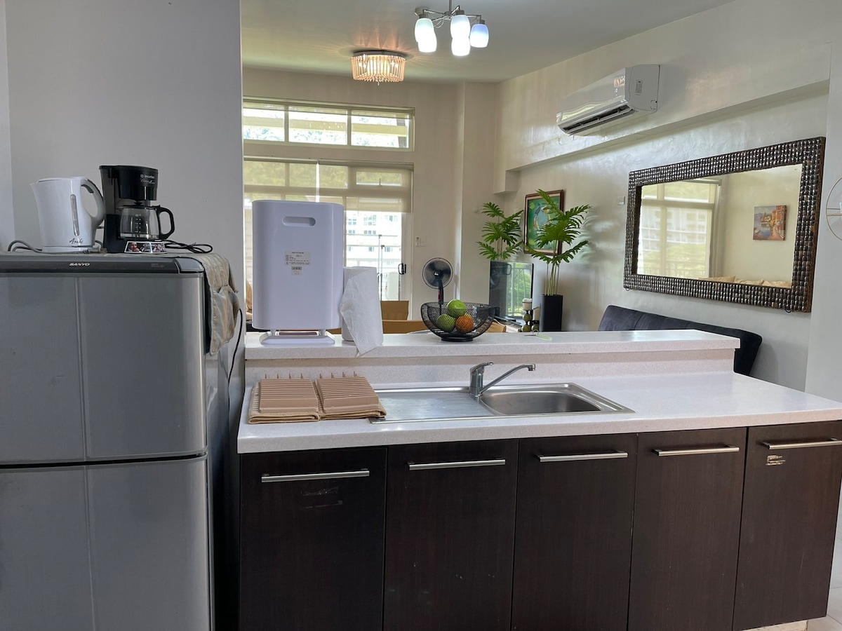 A functional kitchen area is visible, featuring a modern countertop and dark cabinetry. A coffee maker and a refrigerator are positioned along one side. Natural light enters through large windows, illuminating decorative plants and a dining area with a mirrored wall accent.