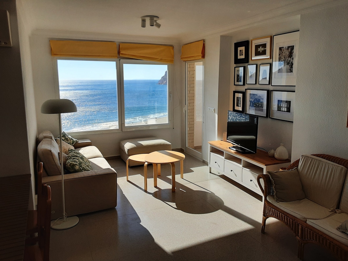 A bright living area is highlighted by large windows that offer expansive views of the sea. A comfortable sofa and wooden coffee table are positioned to take advantage of the natural light. A TV is placed on a side unit, complemented by framed photographs on the walls.