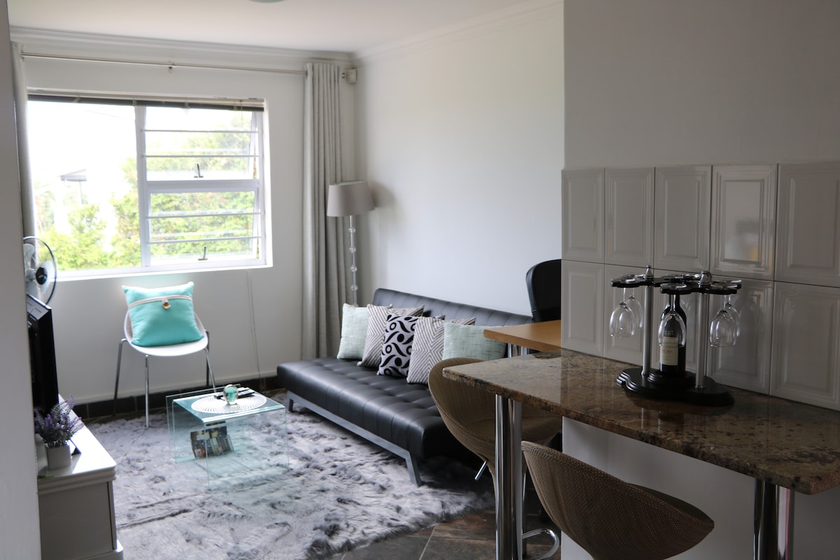 A bright living area features a sleek black sofa with several decorative cushions and a glass coffee table. Natural light enters through a large window, complemented by light-colored curtains. A minimalist bar area is visible, with stylish stools and a granite countertop enhancing the space.