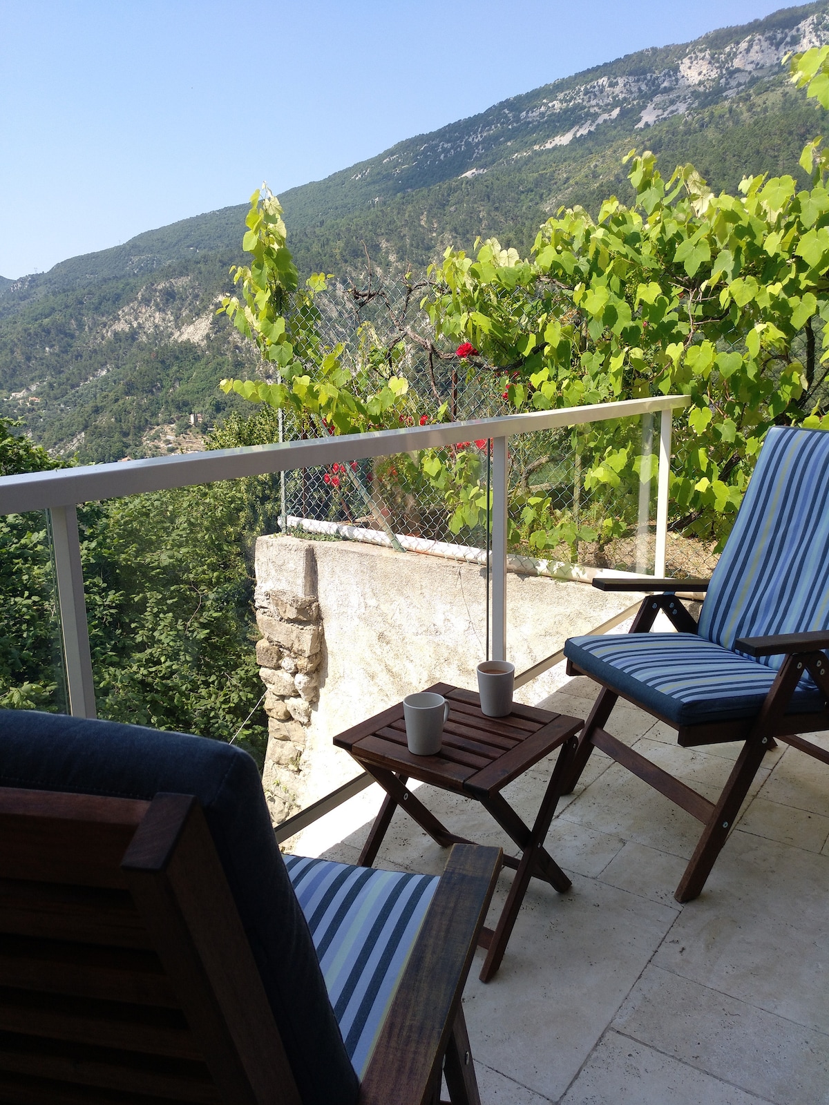 A terrace is visible with two wooden chairs featuring blue and white stripes, positioned beside a small wooden table. Lush greenery and a scenic mountain view create a tranquil setting, enhancing the opportunity for relaxation outdoors.