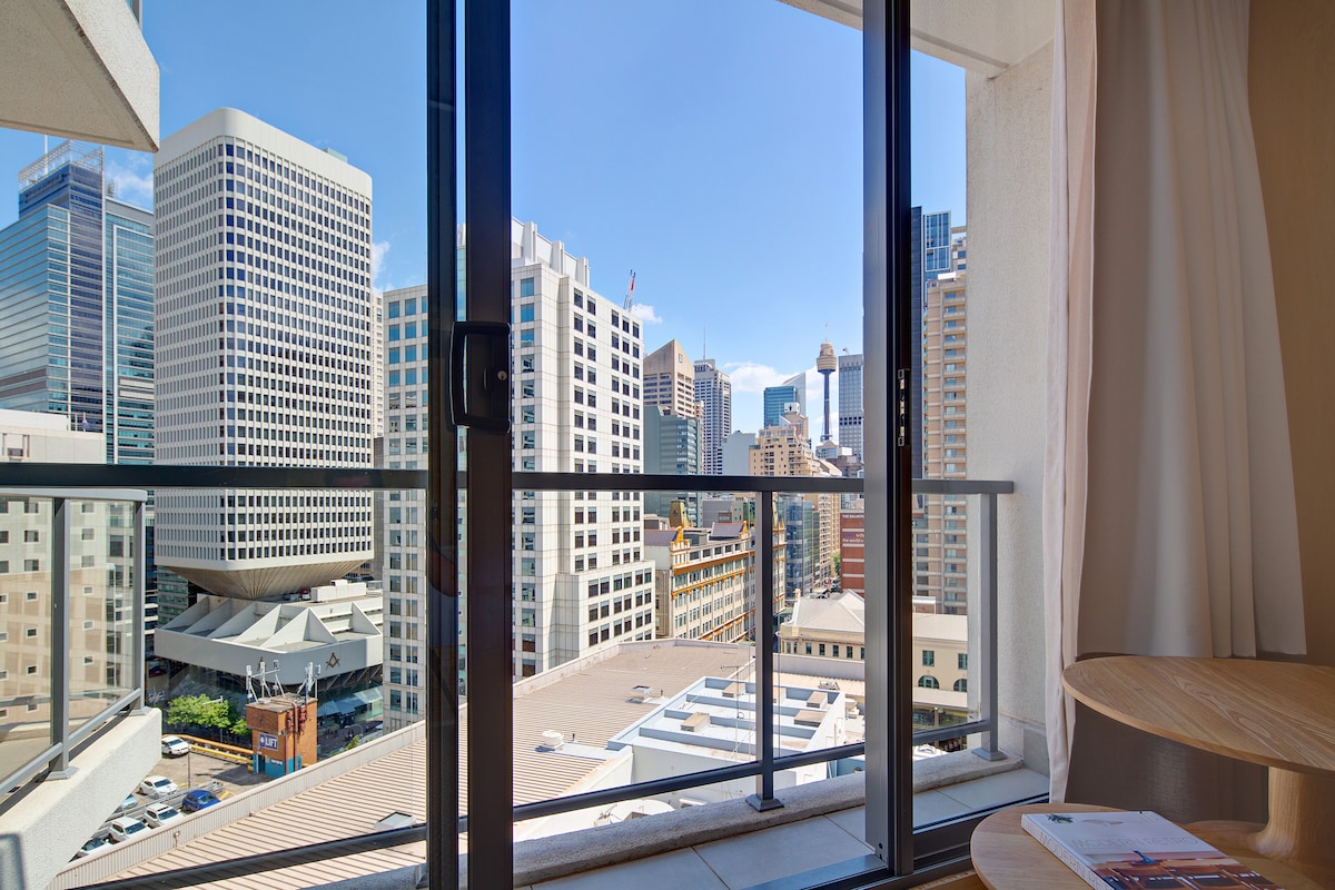 A view from an open-air balcony reveals the vibrant Sydney cityscape. Surrounding buildings reflect modern architecture, with clear skies above creating a bright atmosphere. A small wooden table is positioned nearby, inviting relaxation while enjoying the scenery.